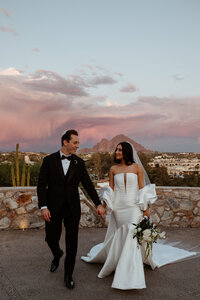 Bride and groom walking hand in hand with a desert mountain and pink evening sky behind them.