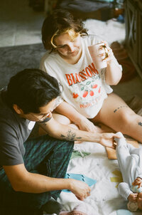 A film photo of a mother and father enjoying their baby laying on a blanket while the mother drinks coffee during their at home family photo session near houston texas