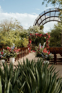 Outdoor wedding ceremony with wooden chairs, colorful aisle flowers and a floral arch surrounded by desert gardens.