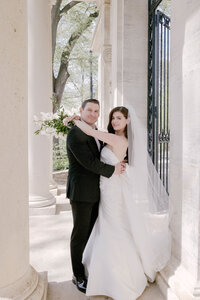 Bride and groom embrace beneath a stone colonnade with an iron gate beside them