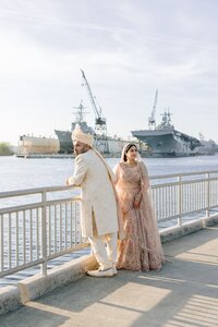 South Asian bride and groom in ornate lehenga and sherwani pose at a sunlit Norfolk harbor railing with a naval ship behind them