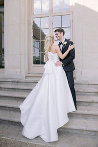 Bride and groom share a smiling embrace on stone steps, her satin bow dress detail visible