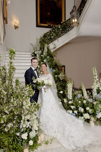 Bride and groom pose on an indoor staircase dressed with a cascading white and green floral garland