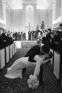 Groom dips bride into a passionate kiss along the chapel aisle as cheering guests applaud in joyful black-and-white elegance