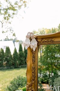 Ivory bow-detailed bridal heels resting atop an ornate gold welcome mirror surrounded by lush garden greenery