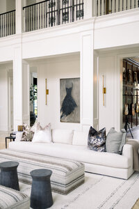 Elevated white living room with plush sofa, patterned pillows, striped ottoman, dark stools, abstract artwork, balcony railings, and guitars.