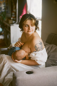 A film photo of a mother nursing her baby in their home and gazing out the window during their at home family photo session near houston texas