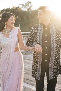 Close view of a couple holding hands at golden hour, the smiling bride in a blush outfit with jewelry and the groom in an intricately embroidered sherwani, at a Hyatt Chesapeake Bay Sikh wedding sangeet