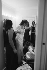 Bride in elegant white gown gets last-minute help from bridesmaids in a warmly lit bridal suite before her intimate chapel wedding