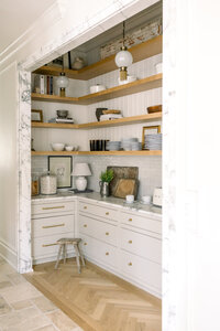 Bright butler’s pantry with open wood shelves, stacked dishes, white cabinetry, pendant lights, and a cozy, elegant feel designed by Buss Studio