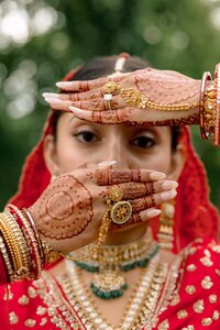 Indian bride in red lehenga frames her face with intricately hennaed hands adorned with gold maang tikka and bangles outdoors