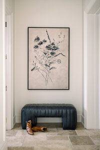Serene hallway with a framed botanical sketch, navy tufted bench, brown boots, and neutral stone flooring, creating a calm, elegant mood.