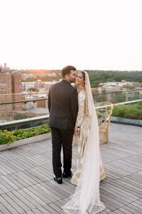 Groom kissing smiling bride in gold-embroidered white bridal gown on a rooftop terrace overlooking Washington DC at sunset
