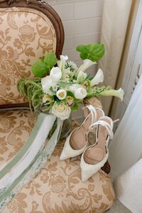 White bridal heels and a lush green-and-white bouquet rest on an ornate vintage chair in an elegant getting-ready suite