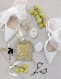 Elegant wedding flat lay with white bridal heels, a vintage gold tray with jewelry, grapes, perfume, and a string of pearls on a soft background.