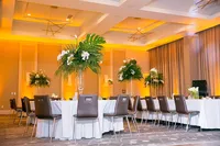 Tables with white tablecloths, brown wooden chairs and tall floral arrangements with lush greenery, palm fronds, roses and orchids