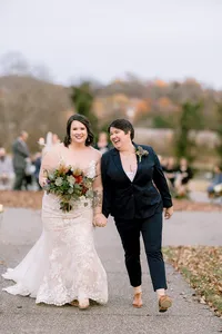 A bride wearing a white lace dress holding a bouquet of yellow, red and orange flowers walks hand in hand with her wife who is wearing a navy suit and small boutonniere of greenery