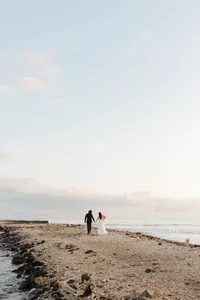 Bride and groom walking hand in hand along the beach during a romantic destination wedding at sunset.