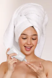 Person with towel wrapped around their head, holding a white bottle and putting white substance from bottle onto their fingertips