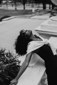 Black and white image with human arched back with sunglasses on, looking up at the sky, with black dress and white sweater wrapped around their shoulders