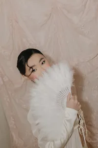 Individual holding white feather fan in front of their face with a beautiful silk background behind them