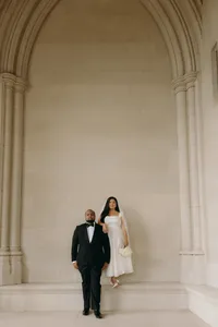 Two humans, dressed in a tuxedo and bridal gown, standing next to one another in front of large pillars.