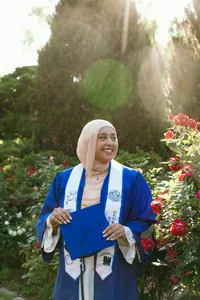 Human in blue graduation gown and holding blue graduation cap, with greenery and flowers blooming around them