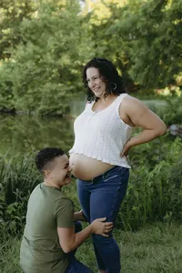 Two humans in outdoor greenery, while one has their face near baby bump, and the other is wearing cropped white shirt