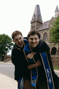 Two humans with graduation sashes hugging one another and looking at the camera with big smiles