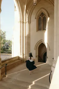 Graduate walking away from the camera to arched doorway with graduation hat on, white top and black gown