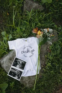Sonogram image, greenery and flowers with a white shirt that says "the little embryo that could"