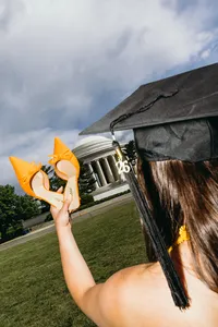 Graduate holding orange shoes in front of building while wearing graduation cap