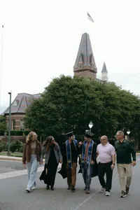 Six humans, with two with graduation hats and sashes, standing in front of greenery and walking towards graduation