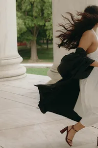 Person in motion walking away from camera with white dress and black gown behind them while dark hair blows in the wind