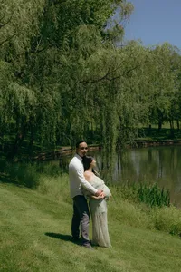 Two humans with one embracing the other from behind and cradling the other's baby bump, both standing in beautiful greenery