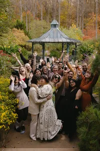 Couple kissing outside at celebration of love while a group of wedding attendees cheer and hold hands up in the background