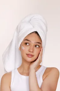 Person with lotion on face and hand on their face with a white tank top on and a white towel on their head looking off to the right