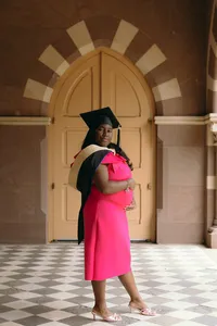 Human standing in hot pink dress with graduation cap on and standing on a brown and white checkered floor with a yellow archway door behind them