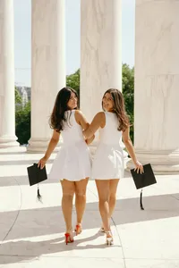 Two humans in white dresses holding graduation caps and looking back at camera