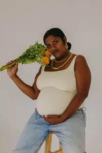 Human holding flowers near face while cradling baby bump in their hand and looking at camera