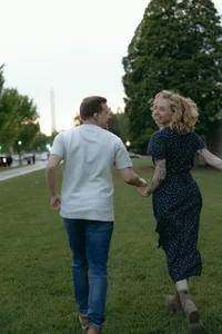 Couple walking outside and holding hands in the green grass, while one looks back at the camera