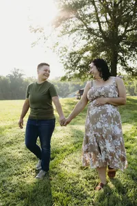 Two humans smiling and holding hands outside in green field, while one holds hand over baby bump