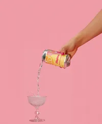 Hand holding colorful can of Anytime and pouring it into a cocktail glass with a pink background