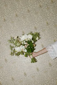 Arm extended and holding bouquet of greenery and white flowers in front of a decorative white and beige background