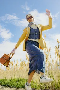 Human in a wheat field with blue sky, while wearing denim outfit, yellow sweater and holding brown bag