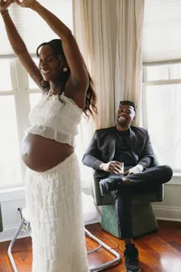 Two humans both smiling, while one is seated in formalwear and the other has their hands up with a baby bump and a cropped white lace shirt and skirt