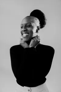 Black and white image of human in black shirt smiling and looking away from the camera, with hands up by their neck.