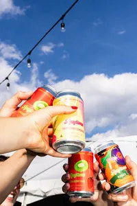 Multiple hands holding up cans of Anytime with string lights above the drinks and blue sky and puffy clouds