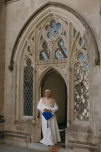Human standing in front of stained glass windows and beautiful university building with white dress and holding blue graduation cap