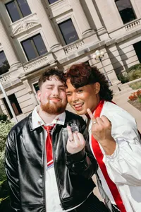 Two individuals looking at the camera and holding up ring fingers with smiles